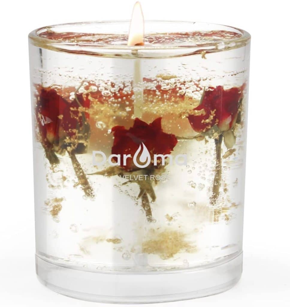 A transparent candle with embedded red roses burns brightly, showcasing the elegance of candles with dried flowers. The words "Daruma Velvet Rose" are printed on the glass, and bubbles surround the roses within the wax.